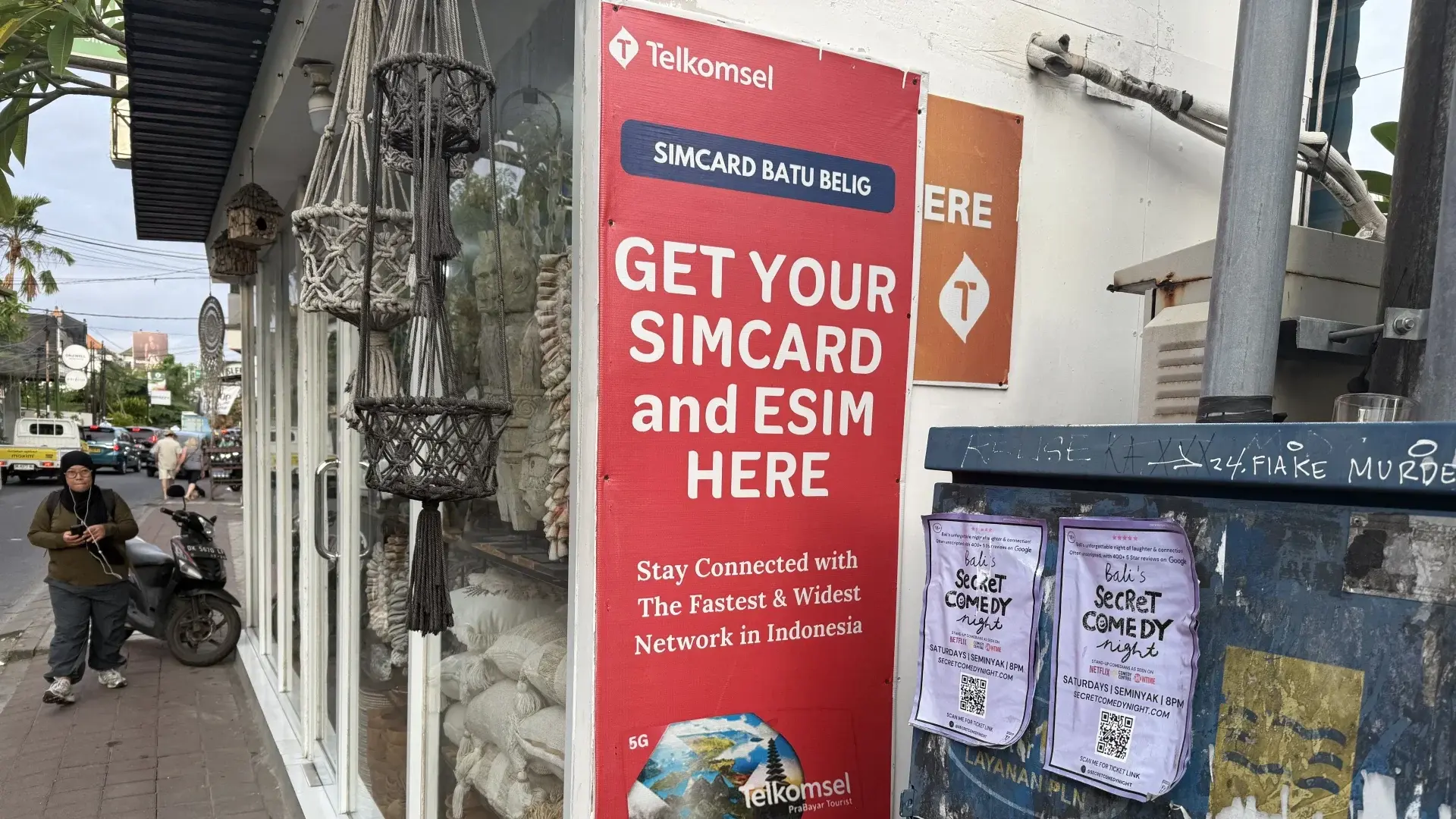 Telkomsel SIM card and eSIM advertisement on a red banner outside a small shop in Batu Belig, near Seminyak, Bali, with a woman walking by and a scooter parked.