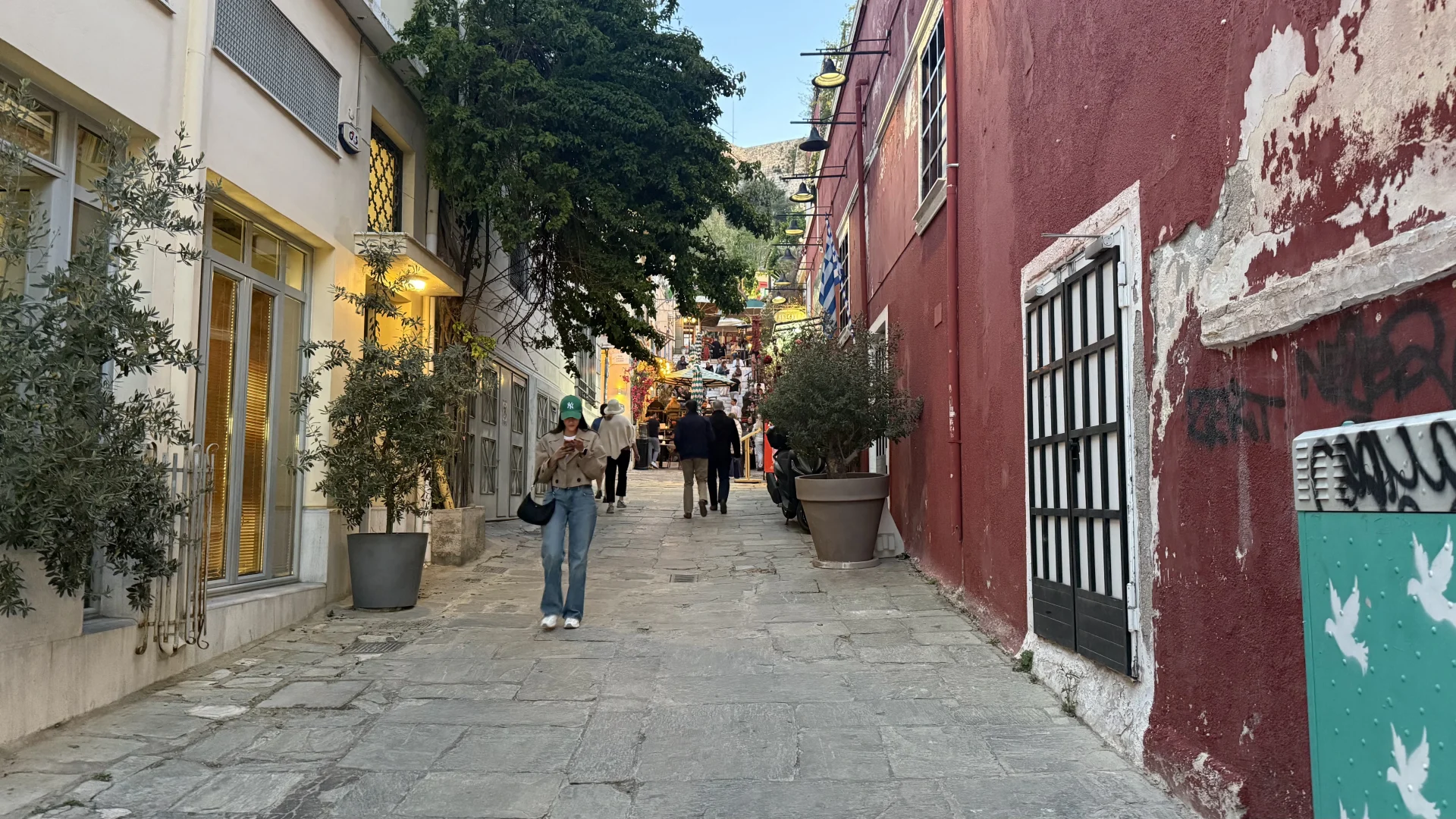 A charming, cobblestone sloping street in the Plaka district of Athens. On the left is a light-colored building with large windows and potted olive trees; on the right, a distinctive weathered dark red wall with graffiti. Pedestrians are walking uphill toward outdoor cafes and lush green trees under a clear sky.