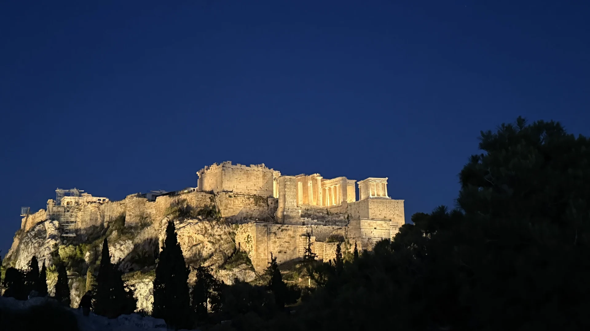 The Acropolis of Athens at dusk, illuminated against a deep blue sky, seen from Filopappos Hill with cypress trees in the foreground