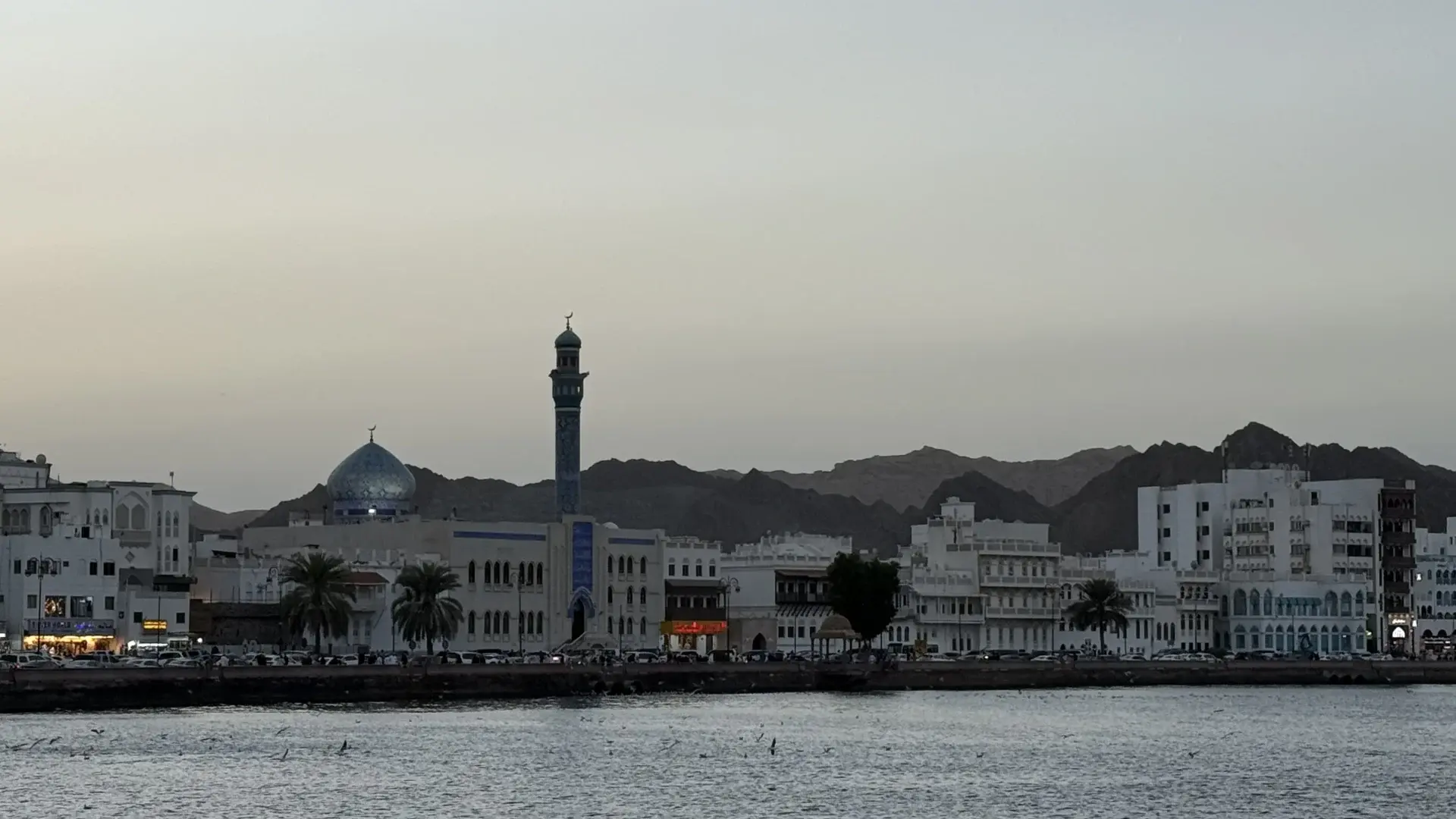 A coastal view of a city in Oman, featuring white buildings, a prominent mosque with a blue dome and minaret, and rugged mountains in the background under a cloudy sky.