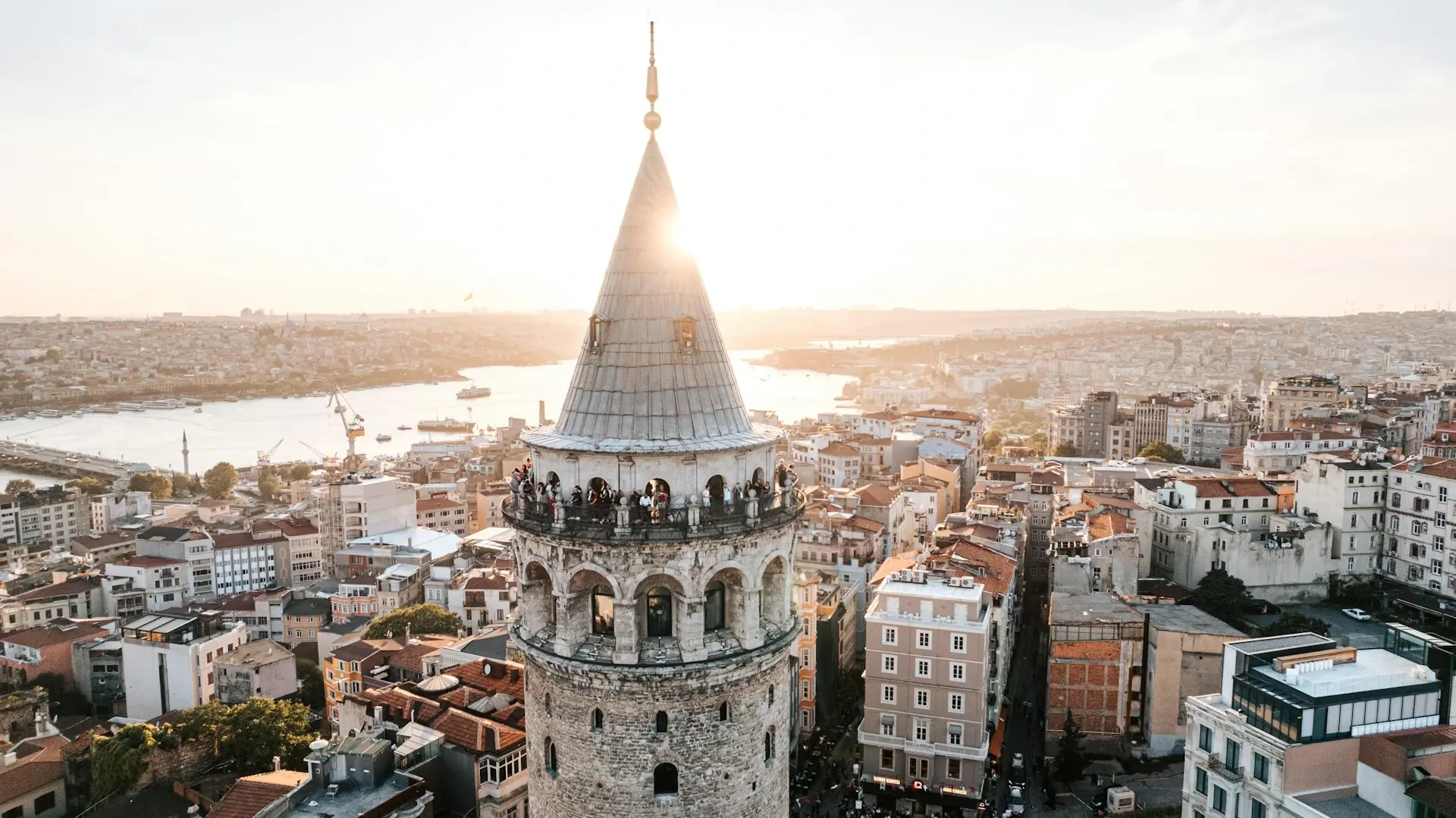 Galata Tower in Istanbul at sunset, with the Golden Horn waterway and the city skyline in the background.