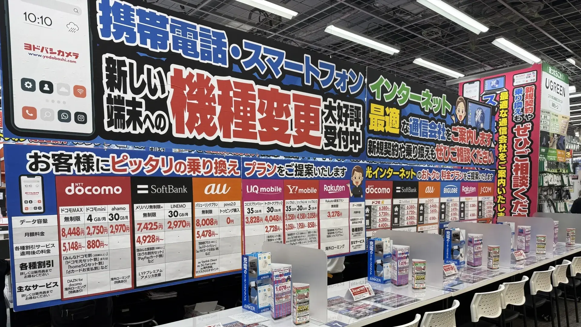 A mobile service counter in a Yodobashi Camera store, displaying large colourful banners and price lists for NTT Docomo, SoftBank, au and Rakuten Mobile