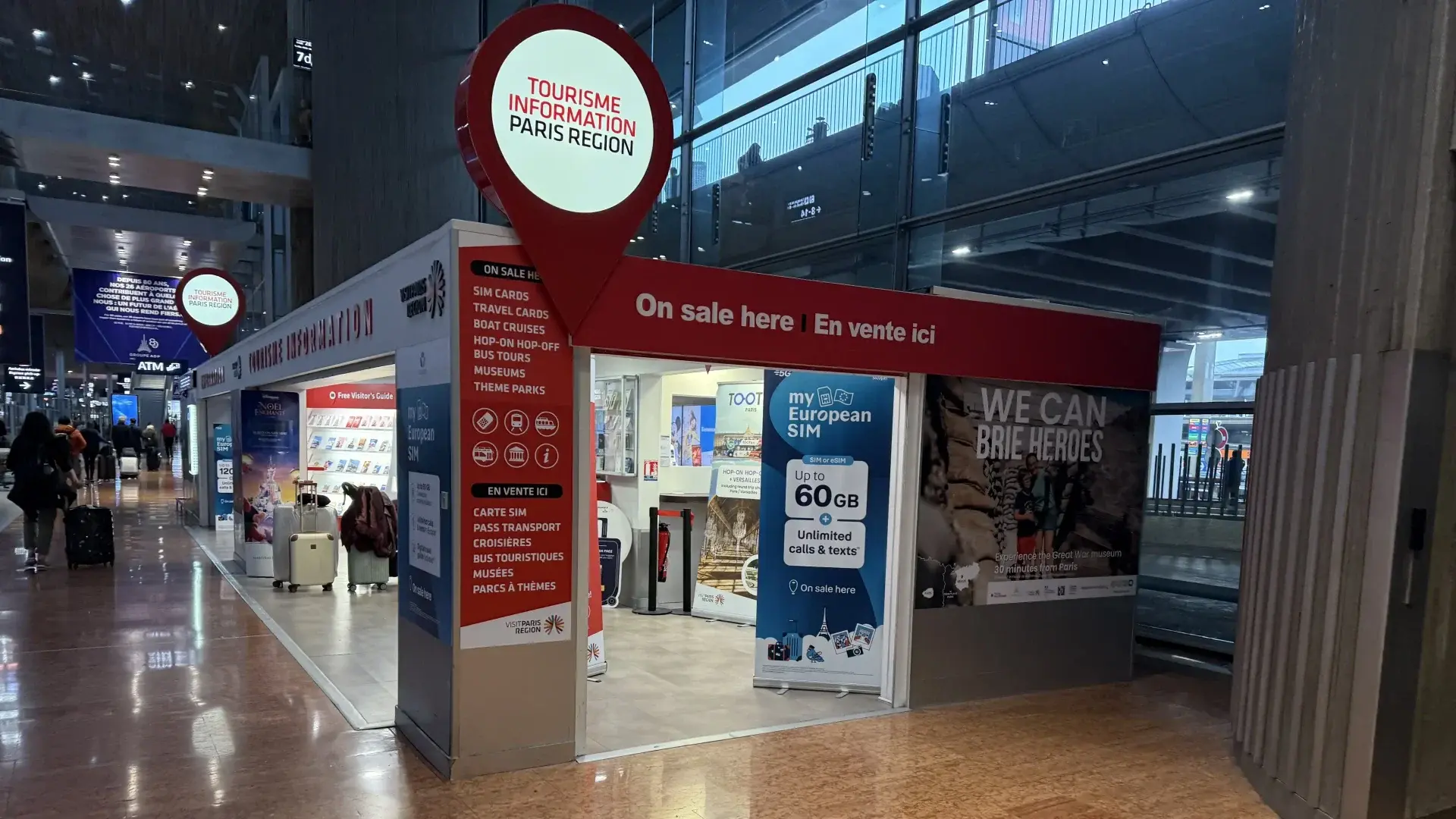 A red and white Paris Region tourism information booth at CDG airport, advertising Bouygues 'my European SIM' cards and travel passes.