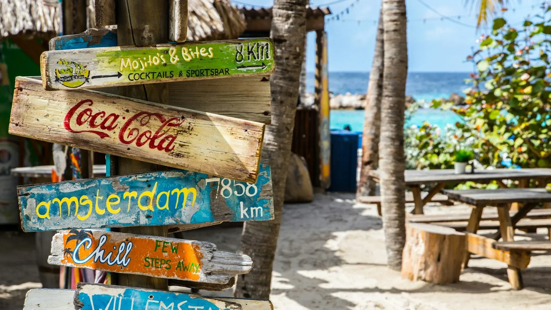 A colourful, rustic wooden signpost on a sunny beach in Curaçao, with painted signs pointing to destinations such as Amsterdam, Willemstad and local beach clubs.