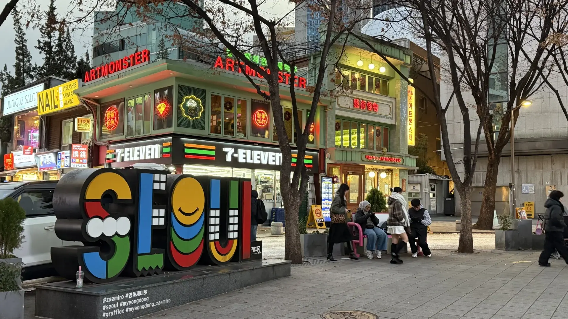 Colourful Seoul street sign in Myeongdong with neon shopfronts, a 7-Eleven convenience store and people sitting and walking in the evening