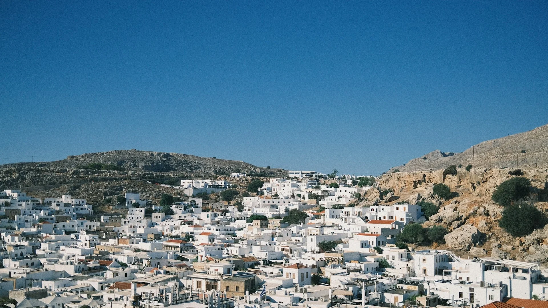 A wide landscape view of Lindos, featuring a dense cluster of traditional white cubic houses built into the side of a dry, rocky hillside under a clear blue sky.