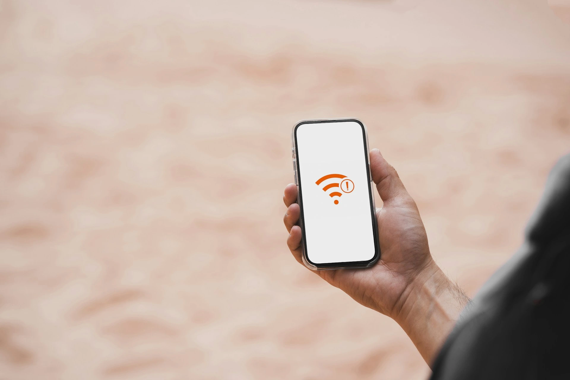 A hand holding a smartphone displaying a Wi-Fi symbol with an exclamation mark, indicating no internet connection or a problem with connectivity. The background is a blurred, light-colored surface.